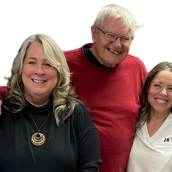 Three people smiling together indoors. On the left, a middle-aged woman with shoulder-length, blonde hair, wearing a black sweater and a gold necklace, smiles warmly. In the center, an older man with short white hair and a red sweater has his arms around both women and is smiling. On the right, a woman in her 30s with light brown shoulder-length hair, wearing a white nurse’s uniform under a striped cardigan, smiles brightly.