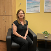 A woman sits in a black armchair in an office-like room with yellow walls. Two framed photos of an ocean scene hang on the wall behind. To the left, there is a wooden cabinet, and to the right, a small table holds a potted plant. A patterned rug covers part of the floor.