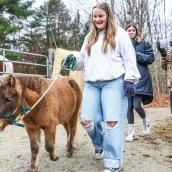 A smiling young woman walks a small pony. Two other young women are in the background. The scene is set outdoors with trees visible in the background. 