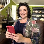 Woman sitting on a couch at home holding a smartphone and smiling, with a cat tree and home décor in the background, suggesting she is comfortably checking information on a mobile app.