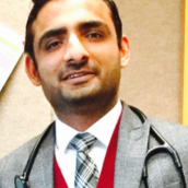 Photo of a man with short dark hair, smiling. He is wearing a grey sports jacket and a red vest underneath with a white collared shirt and a black and white striped tie.