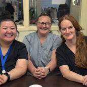 Three women sit together on a couch wearing t-shirts, smiling at the camera.