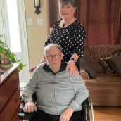 Older couple in their living room. The woman is standing behind her husband with her hand on his shoulder. The man is sitting in a wheelchair.
