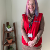 A woman with pink and blue hair stands in a room wearing a red vest next to shelf with plants on it, smiling at the camera.