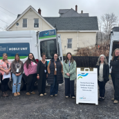 Image of 9 women standing in front of a van that says Nova Scotia Health mobile unit outside. There is a sign propped in between the 7th and 8th woman that says "The Northern Zone Mobile Health and Wellness Team is here on site."