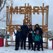 Family with three kids, dad, mom standing in the snow in front of a sign that says "merry"