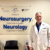 Man in a white lab coat stands in front of a sign on a wall inside a healthcare facility that says "Neurosurgery" and "Neurology".
