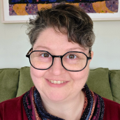 Close up image of a woman with dark curly short hair, wearing glasses and smiling. She is wearing a red top and multicolour scarf.