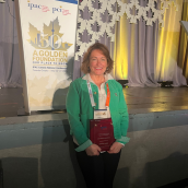 A woman with blonde short hair is wearing a green sweater is smiling at the camera holding an award.