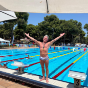 Photo of a man wearing swimming trunks standing at the end of a pool with his arms opened wide. He is wearing a medal around his neck.