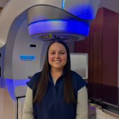 woman with long dark hair standing in front of a radiation therapy machine.