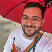 Person with short hair, glasses and wearing white shirt holding a rainbow flag and standing under a red umbrella, smiling at the camera.