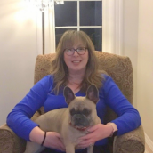A woman sits in a chair inside wearing a blue shirt and is holding a pug dog.