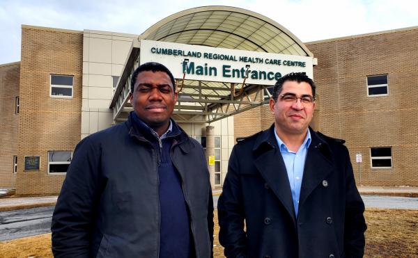 Two men stand in front of a healthcare facility. They are both looking at the camera and wearing blue shirts and black jackets.