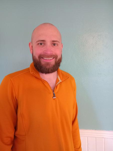 A man with a bald head and full beard smiles at the camera. He is wearing an orange zip-up sweater and standing in front of a light teal wall.