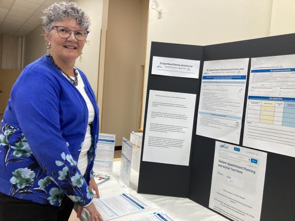 Nancy McBay stands beside a display table featuring the Patient Appointment Planning and Action Tool. She is smiling and wearing a blue cardigan with floral patterns. The display includes posters, handouts, and information about the new tool, which helps patients prepare for healthcare appointments.