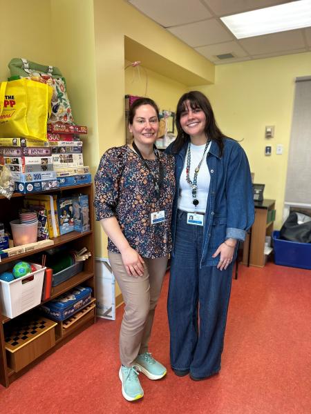 Joy Meloro and Darcy Gould smile together in a brightly lit recreation therapy room at All Saints Springhill Hospital. Shelves beside them are filled with puzzles, games and activity supplies used in patient programs.