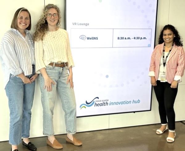 A group of women is standing in front of a large screen. The photo captures them smiling and dressed casually in jeans and footwear. The setting is indoors, emphasizing a collaborative atmosphere.