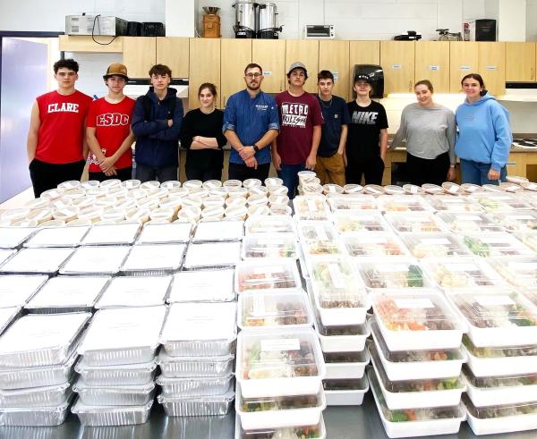 A group of people are standing indoors in front of a large table filled with plastic containers.
