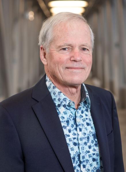 A man with a blue patterned shirt and a navy blue blazer and white hair smiles at the camera.