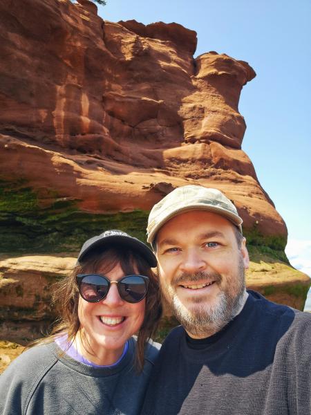Woman with a hat and sunglasses wearing a black sweatshirt is standing beside a man with a beard and moustache wearing a hat and black sweatshirt. They are standing in front of a tall rock formation outdoors, smiling at the camera.