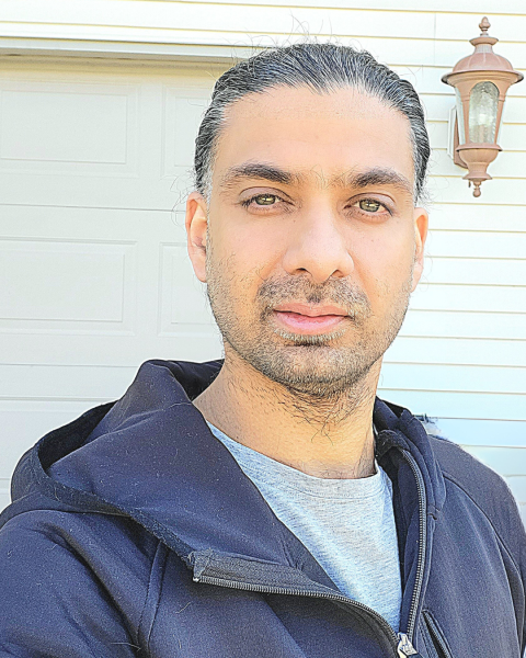 A photo of a man with black hair and beard, wearing a black hoodie over a grey shirt. He is standing in front of a house.