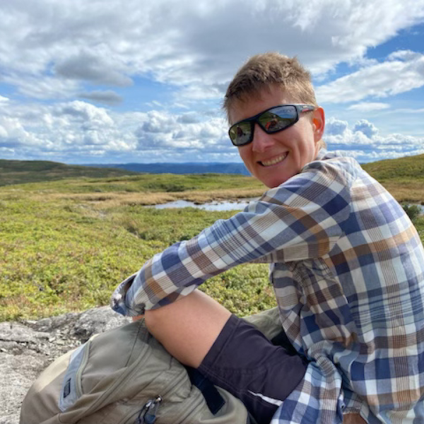 Person wearing sunglasses, a plaid shirt, and shorts sitting on a rocky surface in a grassy, open landscape with small ponds and distant hills under a partly cloudy blue sky.