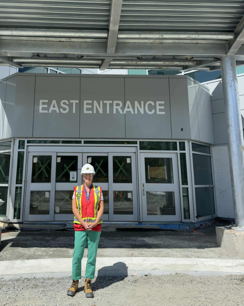 A woman wearing a white hardhat, safety vest, green pants and work boots stands in front of a set of doors on a building with the words "East Entrance" above them.