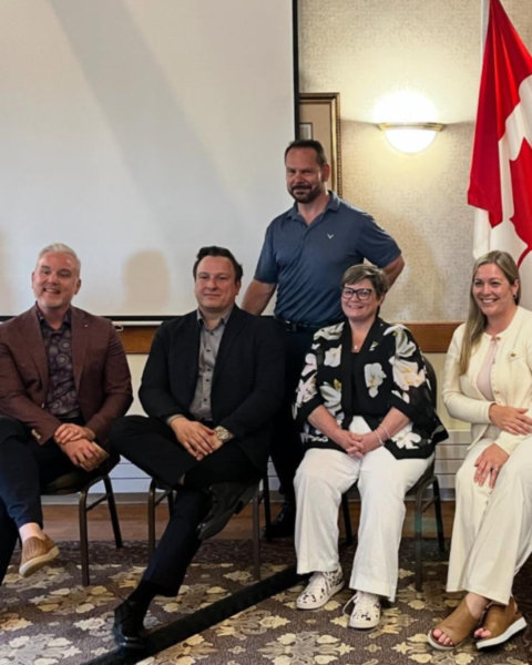 A group of six people are posing for a photo indoors. Four individuals are seated in the front row, while two stand behind them. The setting appears to be a formal or semi-formal event, with a Canadian flag on the right and a projection screen in the background. The floor features a patterned carpet.