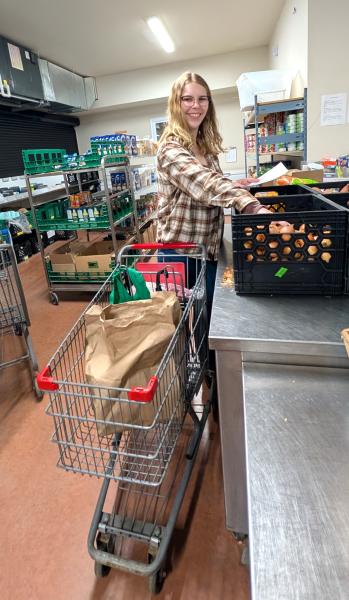 A woman is pushing a shopping cart indoors. The cart is loaded with items and she appears to be actively shopping. The environment includes shelves filled with merchandise and the floor is visible beneath her as she navigates through the space.