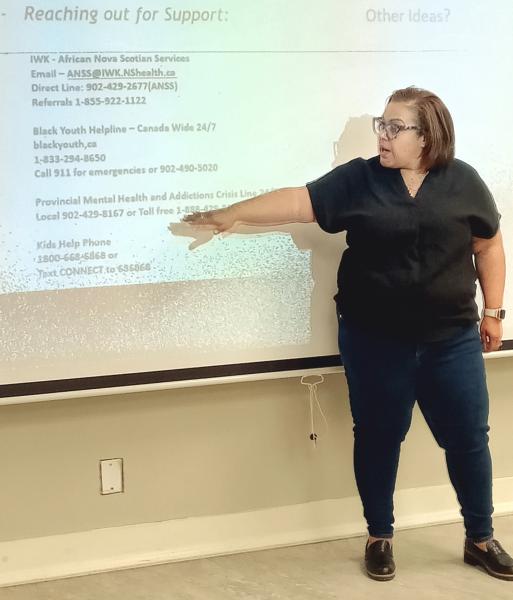 A woman is pointing at a projector screen during a presentation. The content appears to focus on reaching out for support, featuring contact information for various mental health and youth services in Canada. The setting is indoors, with the woman dressed casually in jeans and trousers, engaged in sharing important resources.