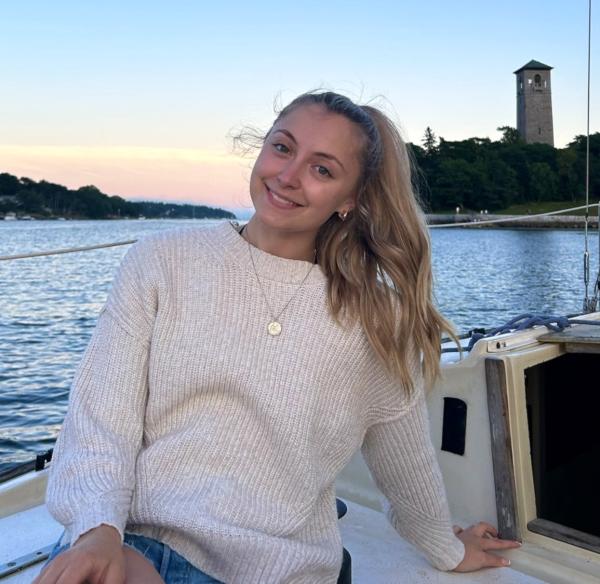 A woman is sitting on a boat, enjoying the outdoors. The setting features clear skies and tranquil waters. She has a smile on her face, dressed in casual clothing.