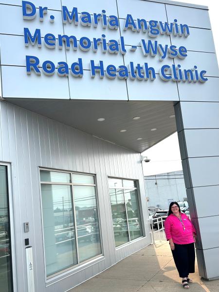 A woman is standing in front of the Dr. Maria Angwin Memorial Wyse Road Health Clinic. The setting is outdoors, featuring a building with a sign that identifies it as the clinic.