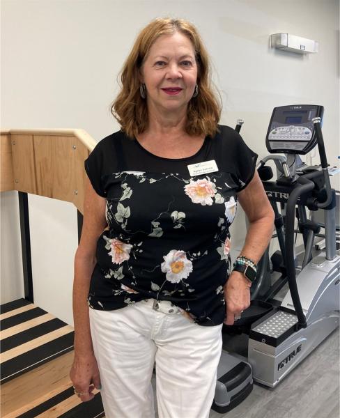 A woman with light, brown hair wears a black shirt, white pants and is standing, smiling at the camera.