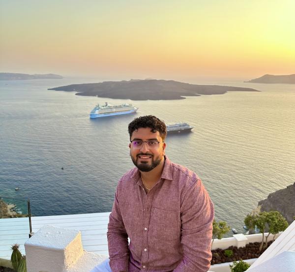 Arjun Sethi, wearing a light red button-up shirt and glasses, sits on a white terrace overlooking the ocean at sunset. Behind him, the calm water reflects the warm glow of the sky, with two cruise ships anchored near a rocky island in the distance.
