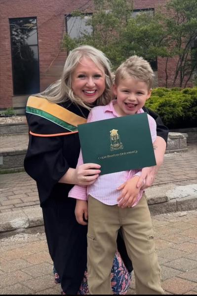 Keshia MacDonald, with long, blond hair, is smiling with her son, also with blond hair, while wearing a graduation gown and holding a diploma.