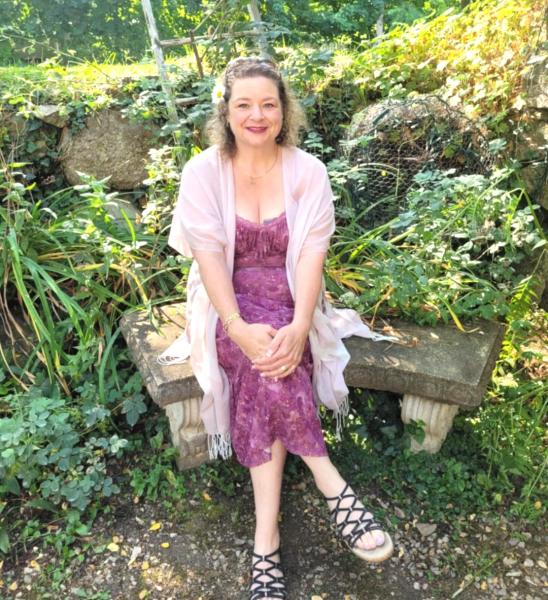 A woman sitting on a bench outside wearing a purple flower pattern dress, highlighting her smile and warm expression. 