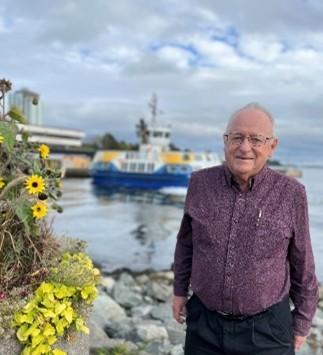 A senior man with gray hair and glasses is wearing a dark purple shirt while standing outside in front of the ocean, rocks and a ferry is in the background.