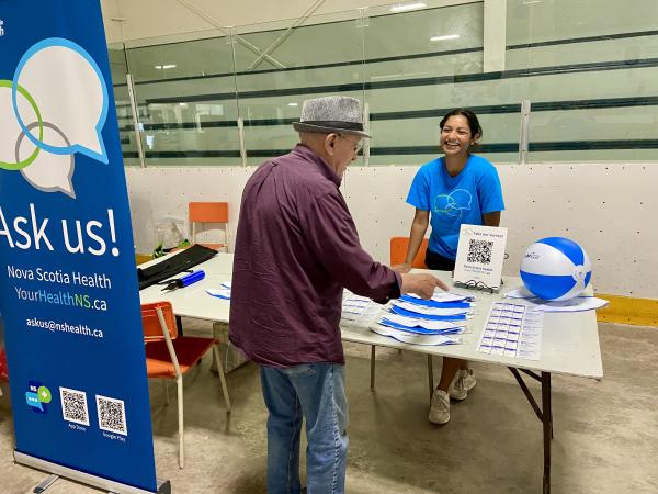 : Roslyn George, Northern Zone Health Ambassador, standing behind an information table with pamphlets, speaking with a community member at a local event.