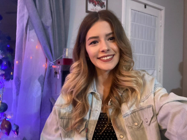 A woman with long, blonde hair is in a bedroom wearing a jean jacket and smiling at the camera.