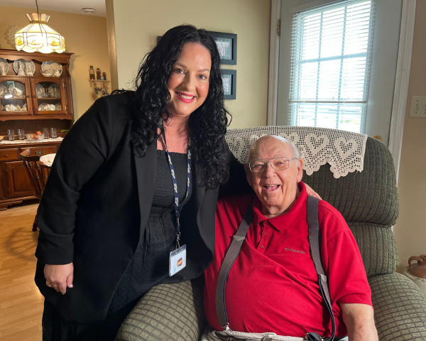 A woman with curly, brown hair and a black shirt and sweater with beige pants sits beside a senior man with a red shirt and suspenders in a living room.