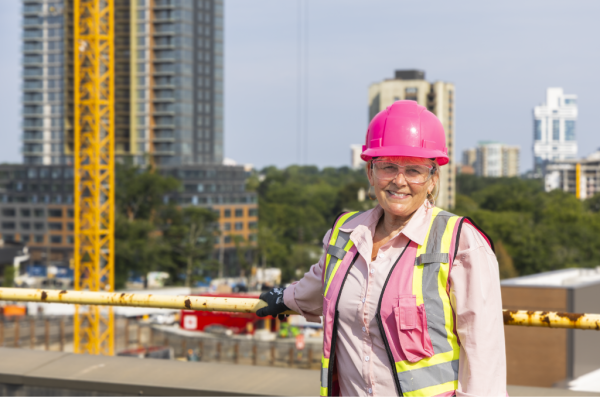 Woman wearing a pink hard hat and safety vest stands with a view of a construction site behind her, smiling at the camera.
