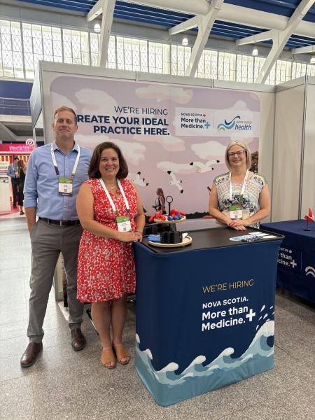 Three Nova Scotia Health representatives stand at a recruitment booth featuring a banner that reads “We’re hiring — Create your ideal practice here. Nova Scotia: More than Medicine.” The group is smiling, and the display table is covered with promotional materials.