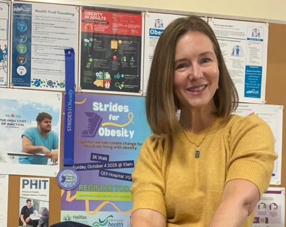 A woman with a dark blonde bob wears a mustard yellow shirt and a pendant necklace while smiling at the camera in front of a bulletin board with colourful posters on it.