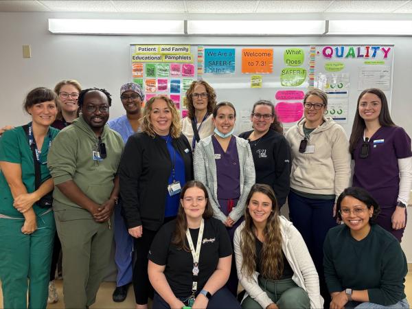 A group of 13 healthcare workers stand together in front of a brightly lit whiteboard, smiling at the camera.