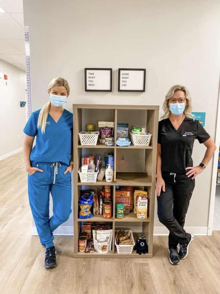 Two women wearing doctor scrubs stand beside a bookshelf full of food and personal care products.