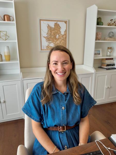 A woman with long brown hair wears a blue dress with a brown belt and is sitting in a room with white cabinets behind her.