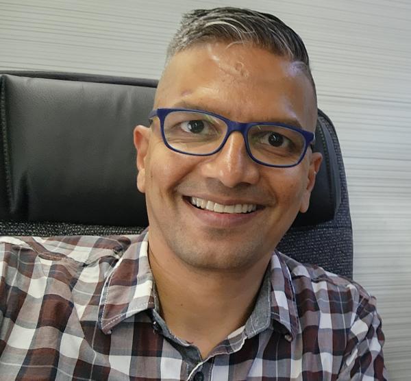 A person with short hair and glasses is smiling while seated in an office chair, wearing a plaid shirt.