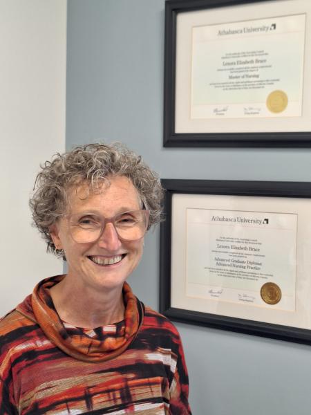 Lenora Brace, nurse practitioner, smiling while standing in front of two framed Athabasca University diplomas on a light blue wall and wearing a striped orange and brown sweater.