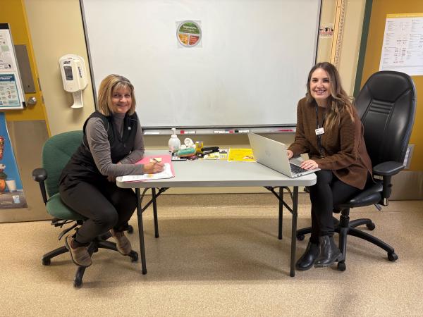 Alice Murphy (left) and Natasha Osmond (right), two Cape Breton diabetes educators, sit at a table in a clinic room. Alice is writing on a clipboard, and Natasha is working on a laptop. Both are smiling toward the camera, with educational materials and medical supplies on the table and a whiteboard behind them.